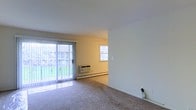 This single-floor residential space offers a straightforward and functional layout centered around an open living area. The main living space is finished with wall-to-wall carpeting that adds warmth underfoot, while clean white ceilings and neutral-toned walls create a bright, airy atmosphere throughout. A full-width sliding glass door floods the room with abundant natural light and provides a direct view of the green outdoor surroundings, offering easy outdoor access. Vertical blinds along the sliding door allow for adjustable light control and privacy. An additional window in the adjoining area further enhances the natural light, keeping the interior feeling open and welcoming. A wall-mounted air conditioning unit and a baseboard heater ensure year-round climate comfort, reflecting practical, all-season functionality. The open flow between the living and adjacent areas makes the space feel connected and versatile, well-suited for a variety of furniture arrangements. Overall, this residence presents a clean, move-in-ready environment with essential amenities in place.