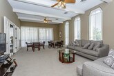 This single-floor space showcases a thoughtfully designed open layout that seamlessly connects a generous living area with a separate dining space. The living room is anchored by exposed ceiling beams that add architectural character and visual height, complemented by a ceiling fan that enhances airflow and comfort. Warm taupe-toned walls provide a neutral backdrop throughout, creating a cohesive and inviting ambiance. Plush, wall-to-wall carpeting in a soft beige tone runs throughout the space, contributing to its comfortable and welcoming feel.

The living area accommodates multiple seating arrangements, with two substantial sofas dressed in patterned accent pillows centered around a round glass-top coffee table. Tall arched windows flood the room with natural light and offer views of the surrounding greenery. A dedicated media corner features a flat-screen television on a multi-tiered media console.

The dining area is bright and airy, featuring two round tables each surrounded by upholstered armchairs, ideal for group gatherings. Sheer floor-length curtains and additional windows with blinds allow for flexible light control. Framed artwork accents the walls, adding a polished finishing touch to this well-appointed space.