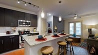 This single-floor space showcases a thoughtfully designed open-concept layout that seamlessly integrates the kitchen and living areas. The kitchen is a standout feature, boasting dark cabinetry that contrasts beautifully with light granite countertops. Stainless steel appliances, including a range and over-the-range microwave, add a contemporary, functional touch. A classic subway tile backsplash lines the cooking area, complementing the overall modern aesthetic. Track lighting runs along the ceiling above the kitchen, while decorative pendant lights hang above the expansive kitchen island, which doubles as a breakfast bar with wooden barstool seating — ideal for casual dining or entertaining. The open layout flows naturally into the living area, where a ceiling fan and natural light from large windows create a bright, airy atmosphere. Warm hardwood-style flooring grounds the space throughout, adding a layer of elegance. Accent walls with subtle textured finishes provide visual interest and depth. The overall ambiance is modern and inviting, balancing sleek design elements with comfortable, livable details that make this a well-appointed and functional space.
