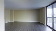 This single-floor space presents a straightforward and functional layout with a clean, neutral aesthetic throughout. The main living area is generously proportioned, providing ample room for a variety of furniture arrangements. The flooring consists of dark hardwood-style planks that run the length of the room, offering a modern contrast against the warm, light beige walls. White baseboards provide a crisp border between the walls and floor, adding a polished finishing detail. The ceiling features a textured popcorn finish and is painted white, contributing to the room's bright and open feel. Electrical outlets are evenly spaced along the walls, ensuring practical accessibility for everyday use. The space is currently vacant, allowing for a clear assessment of its dimensions and potential. The overall ambiance is neutral and versatile, making it well-suited for customization to a range of styles and uses. The combination of dark flooring and light walls creates a balanced visual contrast that gives the space a tidy, well-maintained appearance.