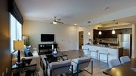 This single-floor residence showcases an open-concept design that connects the living area and kitchen in a cohesive, functional layout. The living space is anchored by a sleek media console with a flat-screen television, complemented by a neutral-toned seating arrangement centered on a geometric patterned area rug. A ceiling fan with an integrated light fixture provides both comfort and ambient lighting, while recessed lighting throughout ensures the space remains well-lit. Hardwood-style flooring extends across the entire area, adding warmth and visual continuity.

The kitchen is positioned at the far end of the open layout and features dark cabinetry contrasted against light countertops and a white peninsula with bar-height seating for two. Pendant lights hang above the peninsula, providing focused task lighting with a stylish touch. A stainless steel sink and modern fixtures complete the kitchen's contemporary aesthetic. A doorway visible from the living area suggests access to additional rooms beyond. Overall, the space presents a clean, modern ambiance suited to everyday living and casual entertaining.