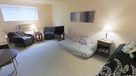 This single-floor space centers around a well-proportioned living area finished with neutral wall-to-wall carpeting that lends a warm, understated feel throughout. The room offers generous floor space, allowing for a comfortable furniture arrangement that includes a patterned daybed-style sofa, a dark upholstered armchair, and a flat-screen television mounted on a compact media stand. Side tables with table lamps are positioned on either side of the seating area, providing soft, ambient lighting that complements the natural light filtering in through a window fitted with horizontal blinds. The walls are painted in a soft, neutral beige tone, creating a clean and calming backdrop. A large abstract canvas in muted blue and grey tones serves as a focal decorative element on the main wall, adding visual interest without overwhelming the space. The overall layout is functional and straightforward, with a clear separation between the seating and viewing areas. The space presents a relaxed, livable ambiance suited for everyday comfort.