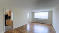 This single-floor space offers a clean, open layout characterized by warm hardwood flooring that runs throughout, reflecting natural light and adding a sense of warmth to the interior. The walls are finished in a soft, neutral tone, creating a bright and versatile backdrop well-suited to a variety of furnishing styles.

The main living area is generously proportioned, with a large picture window dressed in vertical blinds that allows abundant natural light to fill the room while offering views of the surrounding outdoors. A narrow vertical side window near the entryway provides additional light. White baseboards trim the perimeter of the room, complementing the overall clean and understated aesthetic.

A baseboard heating unit runs beneath the main window, providing practical climate control. The smooth, white ceiling contributes to the airy feel of the space. The open floor plan allows for flexible furniture arrangement, making the room adaptable for various living configurations. Overall, the space presents a straightforward, functional design with classic finishes that offer a neutral canvas for personalization.