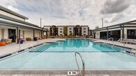 Located at 8651 Airway Dr in Pensacola, Florida, this outdoor amenity space centers around a generously sized swimming pool with crystal-clear turquoise water and classic blue tile accents. The pool features multiple entry points via stainless steel handrails and steps, accommodating swimmers of all ages and abilities. A wide, light-toned paver deck surrounds the pool on all sides, providing ample space for sunbathing and outdoor furnishings such as lounge chairs. On one side of the pool deck, a covered pergola-style structure offers a shaded retreat furnished with teak-framed sofas and chairs, accented by vibrant orange ottomans and blue throw pillows — creating a comfortable, resort-like lounge atmosphere. Ceiling fans are integrated into the covered structure to enhance airflow and comfort during warm Florida days. Lush mature trees and tropical plantings, including palm trees and colorful potted flowers, add natural beauty to the setting. The surrounding residential buildings are visible in the background, providing context for this well-appointed community amenity. The overall design is modern and inviting, blending functional outdoor living with thoughtful aesthetic details suited to the warm Pensacola climate.