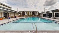 Located at 8651 Airway Dr in Pensacola, Florida, this outdoor amenity space centers around a generously sized swimming pool with crystal-clear turquoise water and classic blue tile accents. The pool features multiple entry points via stainless steel handrails and steps, accommodating swimmers of all ages and abilities. A wide, light-toned paver deck surrounds the pool on all sides, providing ample space for sunbathing and outdoor furnishings such as lounge chairs. On one side of the pool deck, a covered pergola-style structure offers a shaded retreat furnished with teak-framed sofas and chairs, accented by vibrant orange ottomans and blue throw pillows — creating a comfortable, resort-like lounge atmosphere. Ceiling fans are integrated into the covered structure to enhance airflow and comfort during warm Florida days. Lush mature trees and tropical plantings, including palm trees and colorful potted flowers, add natural beauty to the setting. The surrounding residential buildings are visible in the background, providing context for this well-appointed community amenity. The overall design is modern and inviting, blending functional outdoor living with thoughtful aesthetic details suited to the warm Pensacola climate.