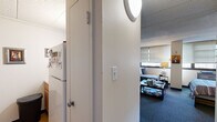 Located at 9 Lamont Ave in Trenton, NJ, this efficient one-bedroom, one-bathroom unit is thoughtfully organized across a single floor. The main living area is generously proportioned relative to the overall footprint, seamlessly blending a living room and sleeping space into one open, functional zone. Large horizontal windows fitted with slatted blinds line the walls, allowing natural light to fill the room while providing privacy. The living area features plush carpeting in a cool blue-gray tone and accommodates a full sofa seating arrangement, a coffee table, a television console, and a dedicated desk workspace — making it suitable for both relaxation and productivity. Framed artwork decorates the walls, adding a personalized touch to the space. The sleeping area is defined by a full-sized bed flanked by a dresser and bookshelf, with a built-in closet providing ample storage. A compact kitchen is tucked efficiently off the hallway and includes a refrigerator and countertop workspace. The bathroom is tiled in classic white with mosaic flooring, and features a wall-mounted sink, toilet, shower with curtain, a porthole-style window for natural light, and an accessibility grab bar. A hallway connects all areas of the unit, completing this well-utilized and practical living space.