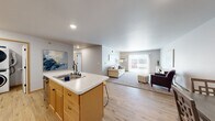 This single-floor unit offers a thoughtfully designed open-concept layout that seamlessly connects the kitchen and living areas. The kitchen is anchored by a central island with a light stone-style countertop, an undermount double sink, and bar seating. Warm-toned wood cabinetry lines the walls alongside stainless steel appliances, including a refrigerator with ice maker, electric range, and over-the-range microwave. Light wood-style flooring runs throughout the kitchen and into the main living space.

The living room is generously proportioned, featuring plush carpet, a wall-mounted AC unit, and a sliding glass door with vertical blinds that fills the space with natural light and provides outdoor access. The neutral gray walls create a calm, cohesive atmosphere throughout.

The primary bedroom is carpeted and includes a wall AC unit, while a second bedroom features a ceiling fan, baseboard heating, and a window admitting natural light. An additional flexible room with carpet, a ceiling fan, and a window serves well as a home office or den. The bathroom is clean and simply appointed with white fixtures and neutral walls.