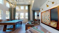 Located at 1079 Beckstead Ct in Denver, NC, this single-story community clubhouse is designed to impress with dramatic double-height ceilings that lend an expansive, airy feel to the main gathering area. A wall of tall, multi-pane windows dressed in bold teal-and-gold geometric-patterned drapes floods the space with natural light while offering views of the surrounding landscape. Two striking gold geometric chandelier pendants serve as focal points overhead, anchoring the room's sophisticated yet playful aesthetic.

At the heart of the space sits a full-size billiards table with a vibrant teal felt surface, complemented by comfortable lounge seating arranged around natural wood accent tables. A shiplap accent wall serves as a dramatic backdrop for an oversized, wall-mounted Scrabble board — a distinctive and interactive design element illuminated by brass swing-arm sconces. Adjacent to this feature wall, a lounge area provides additional casual seating with tasteful artwork.

A dedicated co-working nook features private booth-style seating with upholstered banquettes, rustic wood desks, and geometric pendant lighting — ideal for focused work or small meetings. A kitchenette area with blue cabinetry and a mounted television rounds out the amenities. Wood-look tile flooring in a herringbone pattern ties the entire space together with warmth and style.