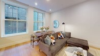 Located at 1401 Spruce Street in the heart of Philadelphia, PA, this single-floor unit offers a sleek, contemporary design with high-quality finishes throughout. Light hardwood flooring runs continuously across the space, complemented by crisp white walls and recessed lighting that create a bright, welcoming ambiance.

The open-concept living and dining area serves as the centerpiece of the unit, featuring large windows that invite natural light and views of the surrounding urban streetscape. A generous sectional sofa anchors the living space alongside a wall-mounted television, while a solid wood dining table with upholstered chairs provides an elegant setting for meals. A smart home control panel is conveniently mounted near the entrance hallway.

The kitchen is outfitted with sleek cabinetry, a striking marble-look quartz countertop and backsplash, an induction cooktop, stainless steel built-in appliances including an oven and microwave, and a deep double sink with a matte black faucet — a refined space suited for everyday cooking.

The bedroom is an intimate retreat featuring a decorative botanical wallpaper accent wall, a transom-style interior window, and warm layered lighting. The bathroom is finished with large-format porcelain tile, a frameless glass walk-in shower, a floating dark vanity with matte black fixtures, and full-height mirrors that enhance the sense of space.