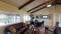 This single-floor space exudes a relaxed, coastal charm defined by its standout architectural feature: a vaulted ceiling with rich, dark-stained exposed wood beams set against a white-painted wood plank ceiling. The open layout connects a comfortable living area to a dining nook, creating a cohesive and inviting atmosphere throughout.

The living area is anchored by a deep brown leather sofa and coordinating armchair, arranged around a dark wood coffee table adorned with a floral centerpiece. A patterned area rug adds warmth underfoot, while a classic table lamp provides soft accent lighting. Framed artwork and photographs line the warm yellow-toned walls, giving the space a personal and welcoming character.

Adjacent to the living area, a round dining table is surrounded by dark upholstered chairs, ideal for intimate gatherings. A tall tropical plant adds a lush, natural element to the corner of the room.

Large windows along one wall flood the interior with natural light and offer views of a lush outdoor area featuring tropical landscaping, including what appears to be a thatched palapa structure, reinforcing the space's laid-back, resort-like ambiance. The overall design blends casual comfort with architectural interest, making it a bright and characterful retreat.