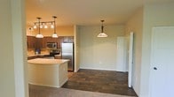 This single-floor space is designed with an open-concept layout that seamlessly connects the kitchen and living areas, creating a fluid and functional environment. The kitchen is a standout feature, equipped with stainless steel appliances including a refrigerator, range, and microwave. Rich, dark wood cabinetry provides ample storage, while a light-toned countertop island serves as both a prep surface and a casual dining or gathering spot. A track lighting fixture with multiple bulbs illuminates the kitchen area, complemented by stylish pendant lights with warm-toned shades that add character and ambiance throughout the space. The flooring transitions from rich, dark hardwood-style planks in the kitchen and dining area to plush carpeting in the adjoining living space, offering both practicality and comfort. The walls are finished in a soft, neutral tone that enhances the brightness of the interior. A hallway visible in the background leads to additional rooms, suggesting a well-organized floor plan. The overall ambiance is warm and inviting, combining modern fixtures with classic design elements to create a comfortable and functional living environment.