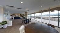This elegant single-floor communal space is designed to offer both comfort and functionality in a well-appointed setting. The open-plan layout is anchored by expansive floor-to-ceiling windows that flood the interior with natural light and provide sweeping panoramic views of the surrounding landscape, including distant mountains and manicured grounds. A large wraparound terrace, accessible through the glazed walls, extends the usable space outdoors and is framed by a sturdy railing system.

Inside, the space is thoughtfully arranged with multiple seating areas to accommodate a variety of gatherings. A casual lounge area features a deep navy sofa and cushioned armchairs, complemented by decorative plants and framed wall art that add a refined touch. Nearby, a more formal dining arrangement includes a round dark-wood table surrounded by upholstered arm chairs in a soft teal tone. A second round table in the foreground further enhances the versatility of the layout.

Underfoot, neutral wall-to-wall carpeting ties the space together, while a textured ceiling with recessed and track lighting ensures even, well-distributed illumination throughout. Structural columns are tastefully integrated into the design, adding architectural character without interrupting the spacious, airy feel of the room.