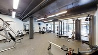 This fitness center offers a comprehensive workout environment designed to accommodate a wide range of exercise needs. The space features an industrial-style ceiling with exposed ductwork and structural columns painted in dark tones, giving the room a modern, urban character. Bright linear LED lighting runs overhead, ensuring the area is well-lit throughout.

The cardio section is outfitted with multiple treadmills and an elliptical machine, all positioned near large floor-to-ceiling windows that allow natural light to filter in and provide views of the surrounding greenery. Wall-mounted flat-screen televisions are positioned above the cardio equipment for entertainment during workouts.

The free weights area includes a rack of dumbbells and is complemented by full-length mirrors along the wall, allowing users to monitor their form. Additional strength training equipment, including weight benches and racks for medicine balls and barbells, is strategically arranged throughout the space.

The flooring consists of durable rubber gym matting in a speckled finish, providing cushioning and traction suitable for a variety of exercises. Overall, the space maintains a functional, well-organized layout that balances equipment variety with open floor space.