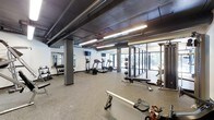 This fitness center offers a comprehensive workout environment designed to accommodate a wide range of exercise needs. The space features an industrial-style ceiling with exposed ductwork and structural columns painted in dark tones, giving the room a modern, urban character. Bright linear LED lighting runs overhead, ensuring the area is well-lit throughout.

The cardio section is outfitted with multiple treadmills and an elliptical machine, all positioned near large floor-to-ceiling windows that allow natural light to filter in and provide views of the surrounding greenery. Wall-mounted flat-screen televisions are positioned above the cardio equipment for entertainment during workouts.

The free weights area includes a rack of dumbbells and is complemented by full-length mirrors along the wall, allowing users to monitor their form. Additional strength training equipment, including weight benches and racks for medicine balls and barbells, is strategically arranged throughout the space.

The flooring consists of durable rubber gym matting in a speckled finish, providing cushioning and traction suitable for a variety of exercises. Overall, the space maintains a functional, well-organized layout that balances equipment variety with open floor space.