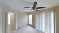 This single-story space in Denton offers a clean, functional layout centered around a comfortable living area. The room features plush wall-to-wall carpeting in a neutral tone, contributing to a warm and understated aesthetic throughout. A five-blade ceiling fan with an integrated light fixture is mounted to the textured popcorn ceiling, providing both air circulation and ambient lighting.

Natural light enters the space through a standard window fitted with horizontal blinds, while a large sliding glass door dressed in full-length vertical blinds serves as the room's primary connection to the outdoors. Together, these openings allow for flexible light control and easy ventilation. The walls are finished in a soft, neutral beige that complements the light-toned carpet and trim.

A doorway visible on the left side of the room leads to an adjacent area, suggesting additional rooms beyond the main living space. White baseboards and door trim provide clean definition against the walls. The overall ambiance is bright, open, and move-in ready, offering a straightforward, practical environment suited to a variety of uses.