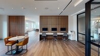 Located at 4307 Sharon Rd in Charlotte, NC, this thoughtfully designed single-floor amenity space blends contemporary elegance with practical functionality. The layout encompasses several distinct zones that flow seamlessly into one another across wide-plank hardwood flooring throughout.

The central lounge area serves as the heart of the space, anchored by a dramatic floor-to-ceiling marble fireplace surround paired with a wall-mounted television. Plush seating arrangements, including tufted sofas and curved accent chairs, create an inviting atmosphere complemented by linear recessed lighting overhead.

A dedicated co-working area features a long white work surface set against a striking vertical wood slat accent wall, accommodating multiple workstations with desktop monitors and task seating. Adjacent to this is a refreshment bar outfitted with sleek white cabinetry, a beverage station, and an embedded display screen above the counter.

The game room offers a poker table beneath a decorative crystal chandelier, framed by warm wood slat partitions that distinguish the space without fully enclosing it. A high-top seating area positioned along expansive floor-to-ceiling windows captures sweeping outdoor views of the Charlotte skyline and treetops, with an adjoining balcony accessible through the glazed facade. Natural light pours throughout, enhancing the space's polished, upscale ambiance.