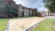 Located at 4500 Highway 57 in Ocean Springs, Mississippi, this outdoor amenity area is a standout feature of a multi-story residential community. The centerpiece of the space is a full sand volleyball court, properly lined and netted, offering residents a dedicated venue for recreational play and social gatherings. The court is bordered by neatly maintained green lawns that provide a clean, open feel to the surroundings.

The backdrop of the space is defined by the community's multi-story buildings, which feature a classic combination of brick and neutral-toned siding, accented by shuttered windows and private balconies. Mature trees and flowering shrubs are thoughtfully placed throughout the grounds, lending a sense of established landscaping and natural shade to the outdoor environment.

The open-air setting benefits from the mild Mississippi Gulf Coast climate, making the volleyball court and surrounding green space enjoyable for outdoor activity throughout much of the year. The layout of the grounds encourages an active and community-oriented lifestyle, with the recreational amenity positioned centrally among the residential buildings for easy access. Overall, the space combines functional outdoor recreation with an inviting, well-kept community atmosphere.