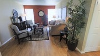 This single-floor space offers a cohesive and well-appointed interior designed for comfortable living. The living area serves as the focal point of the home, showcasing warm hardwood-style flooring that extends throughout and creates a bright, clean foundation for the room. A bold terracotta accent wall adds depth and character, providing a striking contrast against the neutral tones of the surrounding walls and furnishings. Natural light filters in through multiple windows fitted with white blinds, keeping the space feeling open and airy. The living area is furnished with a generously sized tan sofa accented with decorative pillows, complemented by a pair of coordinating armchairs arranged around a circular glass-top coffee table. An ornate area rug anchors the seating arrangement, adding texture and visual warmth. Decorative touches include a large round wall clock, tall potted indoor plants that introduce a natural, organic element, and framed wall art that adds a polished finish. Additional side tables and accent lighting complete the room's layered, inviting ambiance. The textured ceiling adds a classic architectural detail, rounding out a space that balances comfort, style, and practicality in a well-organized single-level layout.