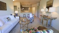 This single-floor residence showcases an open-concept design that integrates the living area and kitchen into one cohesive, flowing space. The living room is anchored by plush neutral seating and a circular glass-and-brass coffee table, accented by boldly patterned multicolor chairs that inject energy and personality into the room. Soft carpet underfoot adds warmth and comfort throughout the space. A console table along one wall provides additional surface space, complemented by warm ambient lighting from table lamps that enhance the inviting atmosphere.

The kitchen is positioned at the rear of the open layout and features dark cabinetry, a light-toned countertop that extends into a raised breakfast bar with bar stool seating — ideal for casual dining or entertaining. Overhead, a track lighting fixture and pendant lights illuminate the kitchen and bar area effectively. The kitchen opens visually to the living space, reinforcing the open-concept flow. A doorway visible from the living area suggests additional rooms beyond. Overall, the space balances a neutral architectural palette with colorful décor for a lively yet comfortable feel.
