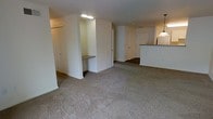 This single-floor residence offers a practical and functional layout designed to maximize everyday living. The main living area is generously sized, featuring wall-to-wall plush carpeting that adds warmth and comfort underfoot. Neutral-toned walls throughout create a clean, versatile backdrop well-suited to a variety of interior styles.

A distinctive built-in desk nook is recessed into the wall, providing a dedicated workspace without occupying additional floor space — a thoughtful architectural detail that enhances the unit's utility. Overhead fluorescent lighting in this area ensures ample illumination for work or study.

The kitchen is positioned adjacent to the living space and features a raised peninsula counter that serves as both a prep surface and a casual dining area. A stylish pendant light hangs above the counter, adding a touch of character to the cooking space. The darker countertop contrasts neatly with the light-colored cabinetry and walls.

A hallway visible from the living area leads to additional rooms, including what appears to be a bedroom or secondary space accessed through a white paneled door. The overall layout flows naturally, with an open sight line connecting the living, dining, and kitchen areas into a cohesive, unified living environment.