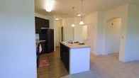 This single-floor residence showcases an open-concept layout that connects the kitchen and living areas in a cohesive, functional design. The kitchen is a standout feature, centered around a large island with a built-in sink and dark cabinetry that contrasts cleanly against the light-toned countertops and neutral walls. Three pendant lights hang above the island, providing focused task lighting while adding a contemporary visual element to the space. Full-size black appliances, including a refrigerator and dishwasher, are seamlessly integrated into the cabinetry layout. The kitchen floor features wood-look hardwood, adding warmth and durability to the cooking area. This transitions naturally into the adjacent living space, where plush carpeting creates a softer, more comfortable atmosphere. Ceiling-mounted lighting throughout ensures the space remains well-lit. A corridor visible beyond the kitchen leads to additional rooms, suggesting a thoughtful and efficient floor plan. The overall ambiance is clean, modern, and functional, with a neutral color palette that provides a versatile backdrop suited to a variety of interior design preferences.