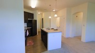 This single-floor residence showcases an open-concept layout that connects the kitchen and living areas in a cohesive, functional design. The kitchen is a standout feature, centered around a large island with a built-in sink and dark cabinetry that contrasts cleanly against the light-toned countertops and neutral walls. Three pendant lights hang above the island, providing focused task lighting while adding a contemporary visual element to the space. Full-size black appliances, including a refrigerator and dishwasher, are seamlessly integrated into the cabinetry layout. The kitchen floor features wood-look hardwood, adding warmth and durability to the cooking area. This transitions naturally into the adjacent living space, where plush carpeting creates a softer, more comfortable atmosphere. Ceiling-mounted lighting throughout ensures the space remains well-lit. A corridor visible beyond the kitchen leads to additional rooms, suggesting a thoughtful and efficient floor plan. The overall ambiance is clean, modern, and functional, with a neutral color palette that provides a versatile backdrop suited to a variety of interior design preferences.