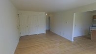 This single-floor space is defined by its clean, understated design and practical layout. The main room is generously proportioned, featuring warm light hardwood floors that extend throughout, lending a bright and inviting character to the interior. The walls are finished in a neutral off-white tone, creating a versatile backdrop suitable for a variety of uses and furnishing styles.

A notable feature of this room is the wall of built-in closets along one side, comprising three separate white panel-door units — two double closets and one single — offering substantial storage capacity. This thoughtful storage arrangement adds considerable functional value to the space.

A flush-mounted ceiling light fixture provides overhead illumination, while electrical outlets are distributed along the walls at standard intervals. A doorway visible toward the rear of the room leads to an additional area, suggesting further rooms or utility spaces beyond what is immediately visible.

The overall ambiance is bright, clean, and neutral — an open canvas well-suited for customization. The space presents itself in vacant, well-maintained condition, with trim and baseboards in good order throughout.