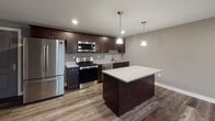This single-floor unit at 2917 Westbrook Dr in Fort Wayne, Indiana, presents a cohesive, modern design highlighted by dark hardwood-style flooring that runs throughout the space. The kitchen serves as a standout feature, equipped with rich dark espresso cabinetry, light granite countertops, and a full suite of stainless steel appliances, including a refrigerator, electric range, built-in microwave, and dishwasher. A central island with a matching granite surface and pendant lighting above adds both functionality and visual appeal, making it an ideal space for cooking and casual dining. The open layout connects the kitchen to the main living area, creating a seamless flow. The bedroom is a well-sized, neutral-toned room with a window that allows natural light to filter in, complemented by the same durable flooring found throughout the unit. The bathroom is finished with a dark vanity topped with granite, neutral taupe walls, wood-look flooring, and a walk-in shower with safety grab bars. Neutral gray and taupe tones are carried consistently throughout the space, lending a clean, contemporary ambiance overall.