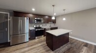 This single-floor unit at 2917 Westbrook Dr in Fort Wayne, Indiana, presents a cohesive, modern design highlighted by dark hardwood-style flooring that runs throughout the space. The kitchen serves as a standout feature, equipped with rich dark espresso cabinetry, light granite countertops, and a full suite of stainless steel appliances, including a refrigerator, electric range, built-in microwave, and dishwasher. A central island with a matching granite surface and pendant lighting above adds both functionality and visual appeal, making it an ideal space for cooking and casual dining. The open layout connects the kitchen to the main living area, creating a seamless flow. The bedroom is a well-sized, neutral-toned room with a window that allows natural light to filter in, complemented by the same durable flooring found throughout the unit. The bathroom is finished with a dark vanity topped with granite, neutral taupe walls, wood-look flooring, and a walk-in shower with safety grab bars. Neutral gray and taupe tones are carried consistently throughout the space, lending a clean, contemporary ambiance overall.