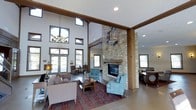 This inviting single-floor space centers around a dramatic great room that immediately commands attention with its soaring two-story ceilings and exposed dark wood beams and columns. The architectural highlight is a floor-to-ceiling stacked stone fireplace, anchoring the room with a rustic yet refined presence and topped with a solid wood mantel. Flanking the fireplace are comfortable seating arrangements, including plush sofas and upholstered armchairs in soft, neutral and teal tones, set atop a rich patterned area rug — creating a warm and welcoming lounge atmosphere. An elegant chandelier hangs near the entrance, complementing a wall of large glass doors and clerestory windows that flood the space with abundant natural light. The high transom windows above the entrance further enhance the sense of volume and openness. Recessed lighting and warm accent lamps add depth and warmth to the interior. The space opens to an adjacent area visible to the right, suggesting additional seating or functional zones that extend the overall layout. The combination of natural stone, wood accents, and soft furnishings creates a cohesive, lodge-inspired aesthetic that is both functional and visually striking.