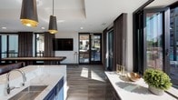 This sophisticated single-floor amenity lounge is thoughtfully designed to accommodate a variety of social occasions. The open-plan layout flows seamlessly across light hardwood and large-format tile flooring, unifying distinct zones that each serve a unique purpose.

At the heart of the space, a generously sized dining and gathering area features multiple round tables paired with tufted upholstered chairs, illuminated by striking gold sputnik chandeliers that add a warm, contemporary flair. A large wall-mounted flat-screen TV anchors one wall, flanked by a white credenza with decorative accents.

A cozy lounge nook offers tufted bench seating set against a bold houndstooth-patterned accent wall, accented by a sculptural gold wall sconce — ideal for intimate conversation or casual relaxation. Adjacent private meeting rooms with glass walls and dark framing provide additional flexibility for smaller gatherings or focused use.

The bar and entertainment area features a navy cabinetry island with a marble-look countertop, a pendant light with a brushed gold interior, and a built-in shuffleboard table nearby. Floor-to-ceiling windows and glass doors dressed with elegant draping curtains flood the space with natural light and open directly to an outdoor terrace, blurring the line between indoor comfort and outdoor living.