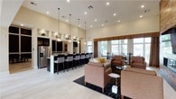 This single-story clubhouse offers a well-appointed, open-concept gathering space designed for both relaxation and socializing. The layout seamlessly integrates a fully equipped kitchen and bar area with a spacious lounge, creating a versatile environment for residents and guests.

The kitchen and bar area features dark cabinetry contrasted by a light-colored countertop, a stainless steel refrigerator, and a row of bar-height seating positioned along the counter. Pendant lighting hangs above the bar, providing focused illumination while adding a modern design element. A textured stone backsplash accent wall enhances the visual interest of this functional space.

Adjacent to the kitchen, the lounge area is furnished with comfortable upholstered armchairs and a sofa in warm neutral tones, accented by decorative throw pillows. The seating arrangements are anchored by a patterned area rug over light tile flooring, contributing to a polished, cohesive aesthetic.

Large windows along the perimeter flood the space with natural light and offer views of the surrounding exterior. The high ceilings throughout add a sense of openness and volume. Recessed lighting complements the pendant fixtures, ensuring the space remains bright and welcoming throughout the day.
