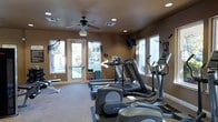 This single-floor fitness center offers a practical and functional workout environment designed to meet a variety of exercise needs. The space is outfitted with a range of cardio equipment, including treadmills, elliptical machines, and a stair climber, all arranged to maximize floor space and ease of movement. A free weight bench and additional accessories are positioned along one wall, providing options for strength training alongside the cardio offerings. Natural light fills the room through multiple large windows and a glass-paneled entry door, creating an open and inviting atmosphere. Recessed lighting and a ceiling fan overhead ensure the space remains well-lit and comfortable during workouts. Warm earth-toned walls give the room a grounded, focused ambiance. A wall-mounted television is positioned for easy viewing from the cardio machines, adding an entertainment element to exercise sessions. The flooring features a low-pile carpet with rubber mat sections beneath the equipment for added stability and floor protection. The overall layout is clean and organized, making this fitness center a convenient and fully functional facility for daily use.