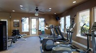 This single-floor fitness center offers a practical and functional workout environment designed to meet a variety of exercise needs. The space is outfitted with a range of cardio equipment, including treadmills, elliptical machines, and a stair climber, all arranged to maximize floor space and ease of movement. A free weight bench and additional accessories are positioned along one wall, providing options for strength training alongside the cardio offerings. Natural light fills the room through multiple large windows and a glass-paneled entry door, creating an open and inviting atmosphere. Recessed lighting and a ceiling fan overhead ensure the space remains well-lit and comfortable during workouts. Warm earth-toned walls give the room a grounded, focused ambiance. A wall-mounted television is positioned for easy viewing from the cardio machines, adding an entertainment element to exercise sessions. The flooring features a low-pile carpet with rubber mat sections beneath the equipment for added stability and floor protection. The overall layout is clean and organized, making this fitness center a convenient and fully functional facility for daily use.