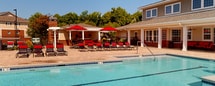 Located at 1000 Broadstone Way in Apex, NC, this outdoor amenity space is designed to offer a resort-style recreational experience. The centerpiece is a generously sized swimming pool with crystal-clear blue water, accessible via wide entry steps and surrounded by an expansive paver deck. Red lounge chairs are arranged throughout the deck area, providing ample space for sunbathing and relaxation under the open North Carolina sky.

Adjacent to the pool, a covered pergola structure serves as a shaded outdoor living and entertainment hub. Beneath it, a built-in brick bar counter with a granite-style top offers a functional gathering space, complemented by red cushioned seating. A ceiling fan overhead ensures comfort during warm days. A dedicated grilling station with a stainless steel grill is positioned nearby, making the area well-suited for outdoor cooking and social gatherings.

The clubhouse building anchors the space architecturally, featuring classic craftsman-style detailing with gray lap siding, brick wainscoting, red accent doors, and white-trimmed windows. A covered front porch equipped with ceiling fans and additional red lounge furniture provides a shaded retreat directly overlooking the pool. The entire area is enclosed by decorative iron fencing, with mature trees lining the perimeter, offering both privacy and a lush, green backdrop.