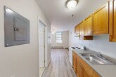 This thoughtfully laid-out single-floor unit makes efficient use of its compact footprint, offering a functional and comfortable living arrangement. The combined kitchen and living area forms the heart of the space, where warm honey-toned wood cabinetry pairs with a stainless steel sink and a white electric range along one wall, while the open living zone benefits from two large windows that invite generous natural light and are fitted with adjustable blinds for privacy. A ceiling fan overhead provides both airflow and ambient lighting. Baseboard heating runs along the perimeter walls, ensuring consistent warmth throughout. Light wood-toned laminate flooring flows seamlessly across the entire unit, lending a clean, cohesive aesthetic. A dedicated closet with classic six-panel double doors provides practical storage. The bathroom is neatly appointed with light tile flooring, a white vanity with under-sink storage, a medicine cabinet with mirror, and a strip light fixture above. Neutral gray and white tones throughout the unit create a bright, airy atmosphere. A short hallway connects the main areas and leads to the entry door, completing this well-organized and move-in-ready space.