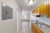 This thoughtfully laid-out single-floor unit makes efficient use of its compact footprint, offering a functional and comfortable living arrangement. The combined kitchen and living area forms the heart of the space, where warm honey-toned wood cabinetry pairs with a stainless steel sink and a white electric range along one wall, while the open living zone benefits from two large windows that invite generous natural light and are fitted with adjustable blinds for privacy. A ceiling fan overhead provides both airflow and ambient lighting. Baseboard heating runs along the perimeter walls, ensuring consistent warmth throughout. Light wood-toned laminate flooring flows seamlessly across the entire unit, lending a clean, cohesive aesthetic. A dedicated closet with classic six-panel double doors provides practical storage. The bathroom is neatly appointed with light tile flooring, a white vanity with under-sink storage, a medicine cabinet with mirror, and a strip light fixture above. Neutral gray and white tones throughout the unit create a bright, airy atmosphere. A short hallway connects the main areas and leads to the entry door, completing this well-organized and move-in-ready space.