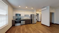 This single-floor unit offers a thoughtfully designed open-concept layout that seamlessly connects the living and kitchen areas. The kitchen is well-equipped with a full suite of stainless steel appliances, including a range, microwave, dishwasher, and refrigerator. Two-toned cabinetry — white upper cabinets paired with dark lower cabinets — frames light granite countertops, creating a clean, modern aesthetic. Wood-look luxury vinyl flooring runs throughout the main living areas, adding warmth and durability to the space. Recessed lighting illuminates the kitchen and common areas, while a ceiling fan provides additional comfort in the living room. Natural light enters through a large sliding glass door fitted with vertical blinds, opening the space to the outdoors. A dedicated in-unit laundry area with a stacked washer and dryer adds everyday convenience. The bathroom features a dark wood vanity with ample storage, a white countertop, and neutral tile flooring, maintaining the consistent modern palette found throughout the unit. A mini-split wall unit provides efficient climate control. Overall, the space presents a clean, contemporary design with practical finishes and a functional, open flow.