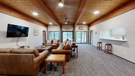 Located at 3675 Highland Ave in White Bear Lake, Minnesota, this single-floor community space offers a thoughtfully designed layout combining a living room and kitchen under one roof. The living room is the architectural centerpiece, showcasing soaring wood-planked ceilings with bold exposed beams that lend a warm, lodge-inspired character to the space. Large floor-to-ceiling windows and French doors flood the room with natural light while offering views to the outdoors. Plush carpeting, leather seating, a wall-mounted television, and a ceiling fan make this an inviting and functional gathering area. A brick accent column adds a rustic touch that complements the overall aesthetic. The adjacent kitchen is practical and well-equipped, featuring gray-toned cabinetry, light stone-patterned countertops, a double-basin sink, a full-size refrigerator, and a microwave. A skylight brightens the kitchen, and a pass-through window connects it directly to the living and dining area, promoting an open, social atmosphere. The dining area includes a round table with seating and bar stools along the counter pass-through, making the space ideal for gatherings and events. Overall, the space exudes a welcoming, rustic-modern ambiance suited for community use.