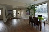This expansive single-floor space is defined by warm hardwood flooring that flows seamlessly throughout, unifying a thoughtfully designed open-plan layout. The main living area is anchored by a dramatic full-wall stone fireplace, crafted from stacked natural stone in earthy tones, flanked by large windows that frame lush outdoor views, including palm trees and a pool. Recessed lighting and a tray ceiling add architectural refinement to the central gathering space, which comfortably accommodates both a seating area and a dining table. A wall-mounted flat-screen TV and built-in speakers enhance the entertainment functionality of this zone. At one end, glass-panel French doors with an arched transom window lead directly to an outdoor patio and pool area, flooding the interior with natural light. A dedicated game room with a billiards table offers a leisure-focused retreat, while an adjacent office or workspace provides functional versatility. The entryway features clean white-framed glass double doors and decorative artwork, creating a welcoming first impression. Lush potted plants are positioned throughout, adding natural vibrancy. The overall ambiance is warm, open, and well-appointed, blending casual comfort with polished architectural details.