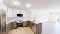 This well-appointed single-floor unit showcases a clean, modern design throughout. The open-concept kitchen is a standout feature, outfitted with two-tone cabinetry — high-gloss white upper cabinets paired with textured wood-grain lower cabinets — set against a classic white subway tile backsplash. Stainless steel appliances, including a French door refrigerator, electric range, built-in microwave, and dishwasher, provide a fully equipped cooking space. A peninsula with a single-basin sink and light countertops extends into the living area, illuminated by sleek pendant lights and flush-mount ceiling fixtures. Light hardwood flooring flows through the main living areas, creating a warm, cohesive feel. The bedrooms are finished with soft wall-to-wall carpeting and feature windows that invite natural light and outdoor views. A sliding glass door opens onto a private balcony, further brightening the living space. The bathroom is finished with large-format marble-look tile flooring, a wood-toned vanity, open built-in shelving, and a walk-in glass-enclosed shower with a rainfall showerhead. Louvered closet doors and recessed lighting add refined, functional detail throughout.