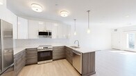 This well-appointed single-floor unit showcases a clean, modern design throughout. The open-concept kitchen is a standout feature, outfitted with two-tone cabinetry — high-gloss white upper cabinets paired with textured wood-grain lower cabinets — set against a classic white subway tile backsplash. Stainless steel appliances, including a French door refrigerator, electric range, built-in microwave, and dishwasher, provide a fully equipped cooking space. A peninsula with a single-basin sink and light countertops extends into the living area, illuminated by sleek pendant lights and flush-mount ceiling fixtures. Light hardwood flooring flows through the main living areas, creating a warm, cohesive feel. The bedrooms are finished with soft wall-to-wall carpeting and feature windows that invite natural light and outdoor views. A sliding glass door opens onto a private balcony, further brightening the living space. The bathroom is finished with large-format marble-look tile flooring, a wood-toned vanity, open built-in shelving, and a walk-in glass-enclosed shower with a rainfall showerhead. Louvered closet doors and recessed lighting add refined, functional detail throughout.