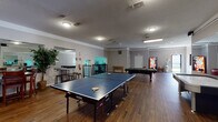This single-floor facility is designed to offer a well-rounded recreational and wellness experience across several distinct areas. The game room serves as a lively centerpiece, featuring hardwood-style flooring, textured ceilings, and ample overhead lighting. It is outfitted with a full-size JOOLA ping-pong table, a pool table, and a vending machine, making it an ideal space for social activity and leisure. A large mirror on one wall visually expands the room, and a seating area with bar-height tables and stools provides a casual spot to relax.

Adjacent to the game room is a lounge and dining area furnished with a leather sofa, a wooden dining table with chairs, and decorative indoor plants that add a welcoming, lived-in feel. The warm hardwood-style flooring continues throughout this zone, creating a cohesive aesthetic.

The fitness center is expansive and divided into functional zones. One area houses cardio equipment including treadmills, elliptical machines, and exercise bikes, complemented by wall-mounted mirrors. A separate strength training area is equipped with cable machines, a power rack, kettlebells, dumbbells, a punching bag, and weight benches, all set on durable rubber flooring. The clean, bright white walls and high ceilings contribute to an open and motivating workout environment.