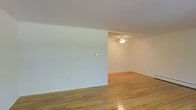This single-story space presents a straightforward, open layout characterized by its clean aesthetic and functional design. The interior is finished with warm-toned hardwood flooring throughout, providing a classic and durable foundation that adds visual warmth to the otherwise neutral palette.

The walls are painted in a crisp white, creating a bright and airy atmosphere that makes the space feel open and adaptable. A partial interior wall divides the main area into two connected zones, offering a subtle sense of separation while maintaining an open flow between spaces. This layout provides flexibility for arranging furniture or defining distinct functional areas.

A ceiling fan is mounted in the secondary zone, contributing to air circulation and adding a modest lighting fixture overhead. The white ceiling is smooth and consistent throughout, complementing the simplicity of the overall design.

Baseboard trim runs along the base of the walls, adding a subtle architectural finish. The space is unfurnished, allowing the structural and finish details to stand out clearly. Overall, the interior offers a clean, neutral canvas well-suited for a variety of uses and personal styling preferences.