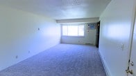 This single-floor space offers a clean, functional layout designed with practicality in mind. The main living area is generously sized, featuring soft wall-to-wall carpeting that adds warmth underfoot and a neutral color palette throughout that creates a bright, open atmosphere. A large picture window allows natural light to fill the room, enhancing its airy feel while offering views of the surrounding exterior. Climate control is well-covered, with a wall-mounted air conditioning unit positioned above the window for cooling and baseboard heating units running along the perimeter walls to ensure warmth during colder months. The textured popcorn ceiling adds a classic touch to the space. The room's open layout provides a versatile canvas, easily adaptable for a variety of furniture arrangements and living configurations. A doorway visible at the far end of the room suggests additional living or utility spaces beyond, contributing to an overall sense of functional flow throughout the unit. The minimalist, neutral design makes this space highly adaptable to a range of personal styles and needs.