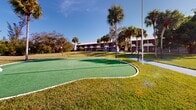 Located at 7235 Maida Ln in Fort Myers, Florida, this well-maintained community outdoor space offers a range of recreational amenities set against a lush tropical backdrop. The grounds are anchored by a multi-hole putting green with a smooth artificial turf surface, complemented by a dedicated shuffleboard court — both ideal for leisurely outdoor activity. Towering sabal palms and mature shade trees are distributed throughout the property, lending a distinctly Florida character to the landscape.

A tranquil pond runs along one side of the grounds, with a paved walkway tracing its edge and offering pleasant waterside views. Stone picnic tables and benches are positioned on concrete pads throughout the property, providing comfortable outdoor gathering spots shaded by the surrounding palm canopy. Manicured green lawns connect each amenity area, with curved pathways guiding residents between them.

The two-story residential buildings frame the perimeter of the grounds, featuring covered walkways and balconies that overlook the common areas. A children's play structure is also visible on the grounds. Globe-style lamp posts are evenly spaced throughout, ensuring the outdoor areas remain accessible and well-lit after dark. The overall atmosphere is serene, social, and quintessentially Floridian.