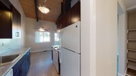 Located at 24797 Joyce St in Hayward, California, this single-floor residence showcases a distinctive architectural character defined by vaulted ceilings with exposed dark wood beams and a natural wood plank ceiling finish. The living area is a standout space, combining generous vertical volume with an abundance of natural light streaming through multiple windows and a sliding glass door that opens to a private balcony, offering outdoor views of the surrounding neighborhood. Soft carpeting and neutral wall tones contribute to a calm, comfortable ambiance throughout.

A brick fireplace with a dark surround serves as a focal point in one of the main living areas, adding warmth and visual interest. The galley-style kitchen is efficiently designed with dark espresso cabinetry extending to ceiling height, granite-style countertops, white appliances including a refrigerator, electric range, and dishwasher, and light wood-tone flooring that transitions cleanly through the space.

Bedrooms feature plush carpeting, neutral walls, and windows providing natural light. At least one bedroom includes a wide sliding-door closet with built-in shelving for ample storage. The bathroom is finished with white tile surrounding a full soaking tub and shower combination, complemented by chrome fixtures in a clean, functional layout.