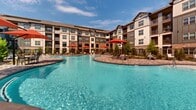 This outdoor amenity space centers on a generously sized, freeform resort-style pool with vivid turquoise water that serves as the focal point of the community's exterior. The pool features a smooth, curved shoreline design with a gradual wading entry point, making it both visually striking and highly functional. Surrounding the water is a broad, patterned paver deck finished in warm earth tones, providing ample space for lounging and circulation. Multiple seating areas are distributed across the deck, furnished with contemporary outdoor chairs and accented by bold red market umbrellas that provide shade throughout the day. Lush landscaping with young trees and ornamental plantings frames the perimeter, softening the hardscape and adding greenery to the environment. A wrought iron fence encloses the space, defining boundaries while preserving open sightlines. The backdrop is formed by a multi-story building constructed with a combination of stone veneer, horizontal siding, and dark wood-toned trim, featuring craftsman-style gabled rooflines and private balconies. The overall ambiance is resort-inspired, combining an upscale aesthetic with a relaxed, open-air atmosphere ideal for outdoor recreation and social gatherings under open skies.