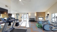 This fitness center is a thoughtfully designed, single-floor facility built to support a well-rounded workout experience. The space features a patterned carpet tile floor in warm tan and cool gray tones, adding a modern, energetic aesthetic underfoot. Large glass doors at the far end flood the room with natural light and provide direct access to an outdoor area, creating an open and inviting atmosphere.

The facility is equipped with a variety of cardio machines, including treadmills and a stationary bike, positioned to take advantage of the natural light. A dedicated free weights station features a fully stocked dumbbell rack, while a green step platform and additional accessories offer versatility for functional training. Numbered workout stations are clearly marked, supporting organized group fitness or circuit training routines.

A striking stacked stone fireplace anchors one wall of the room, offering a warm and distinctive architectural focal point that sets this fitness center apart. Wall sconces provide warm accent lighting alongside the main ceiling fan. Safety essentials, including a fire extinguisher and emergency exit signage, are visibly placed throughout. Overall, the space balances practicality with comfort, making it a functional and welcoming environment for daily exercise.