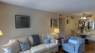This single-floor space presents a comfortable and well-appointed living room as its central feature. The room is finished with warm beige walls and a textured ceiling, contributing to an intimate and relaxed atmosphere. Dark hardwood flooring anchors the space, providing a rich contrast to the light-colored furnishings throughout.

The living area is centered around a cream-colored sofa adorned with decorative gray and blue accent pillows and a soft gray throw blanket, creating a layered, inviting look. A cushioned blue accent chair adds a complementary pop of color and additional seating. A side table with a classic table lamp provides warm ambient lighting, enhancing the cozy feel of the room during evening hours.

Wall-mounted framed artwork in muted tones adds visual interest to the neutral walls without overwhelming the space. A wall clock near the hallway entrance adds a practical decorative touch. Ceiling vents suggest a central HVAC system for year-round climate comfort. A doorway visible in the background hints at additional rooms beyond, indicating a thoughtfully connected single-level floor plan designed for easy, accessible living.